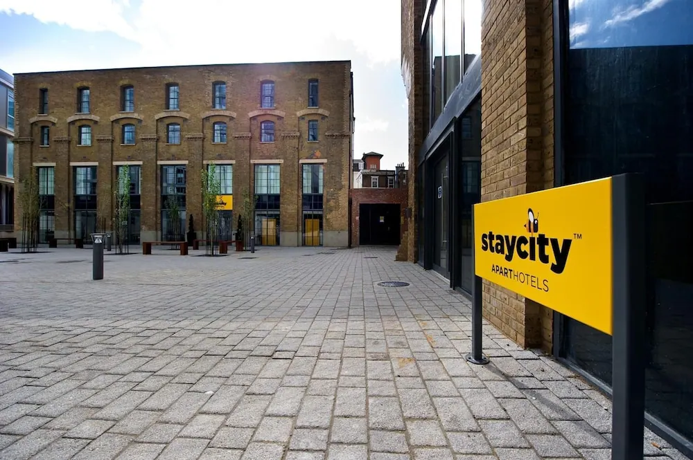 A paved courtyard with a yellow-brick building and a yellow staycity Aparthotels sign on a metal post in the foreground.