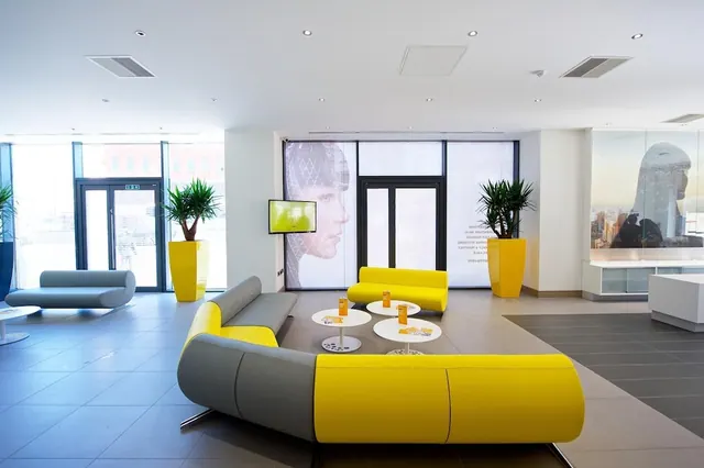 A lobby with yellow and grey sofas, round white tables with orange drinks, potted plants in yellow planters, and a wall-mounted screen beside tall glass doors.