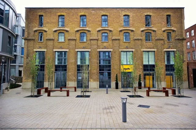 A tall brick building with arched windows stands behind a paved courtyard with wooden benches, young trees and bollard lights.
