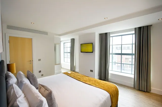 A bedroom with a double bed dressed in white linen and a mustard throw, a wall-mounted television, and large windows overlooking brick buildings.