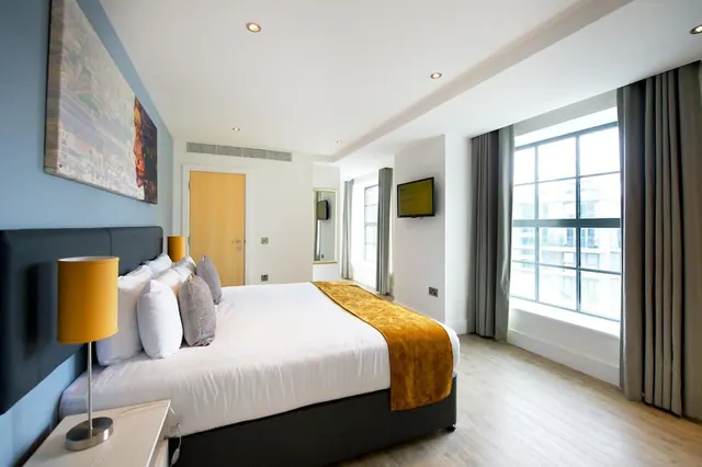A staged double bedroom with a blue accent wall, a wall-mounted television and a large window looking onto a neighbouring building.