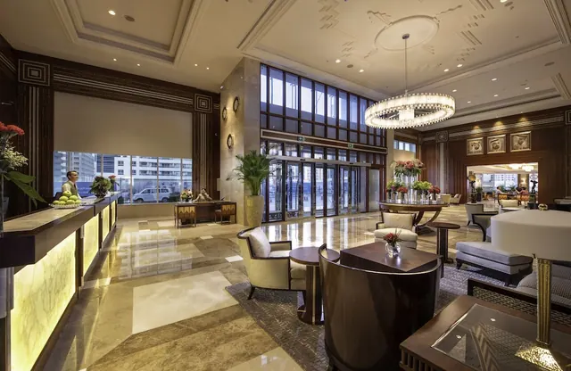 A hotel lobby with a reception desk on the left, a large chandelier overhead, seating groups on a marble floor, and glass entrance doors with a street visible outside.