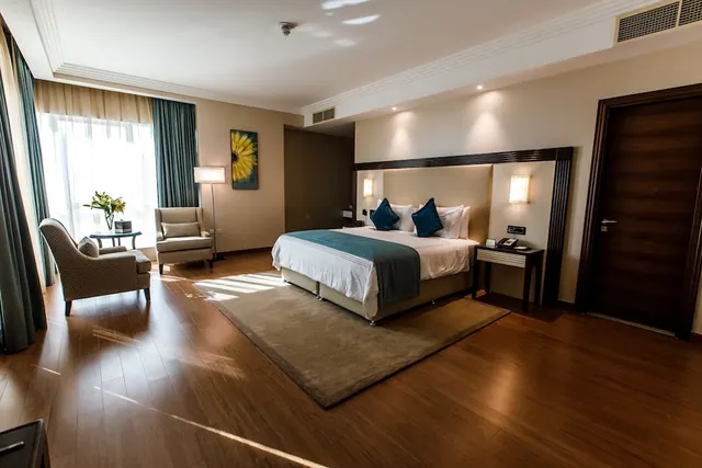 A hotel bedroom with a made double bed with teal cushions and throw, two armchairs beside a window with sheer curtains, and a polished wooden floor.