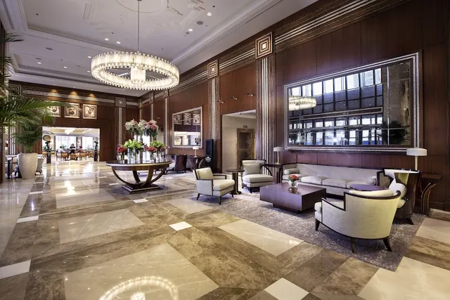 A large hotel lobby with a tiered crystal chandelier, a marble floor, wood-panelled walls, and seating groups around a round table of flowers.