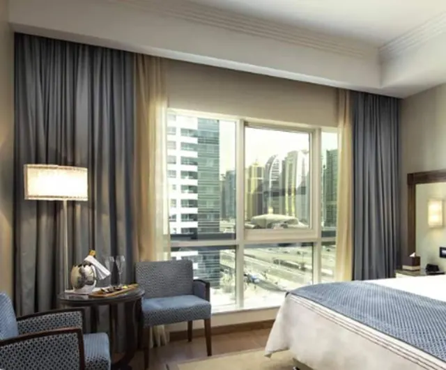 A hotel bedroom with a bed, two armchairs beside a small round table, and a large window overlooking high-rise towers and a road.