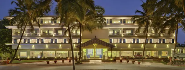 A brightly lit tropical hotel exterior is framed by palm trees at dusk.