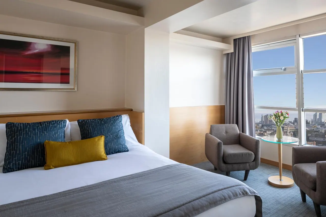 A double bed with blue and yellow cushions sits in a hotel bedroom beside two armchairs and tall windows looking over a city skyline.