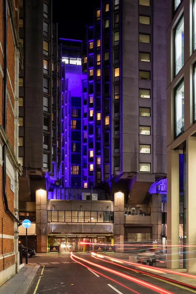 Night view up a street toward a tall hotel building lit with purple uplighting, with car light trails on the road in the foreground.