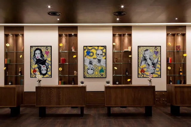 A hotel lobby with wooden reception counters, lit wooden display shelves holding bottles and flowers, and three framed pop-art portraits on a cream wall.