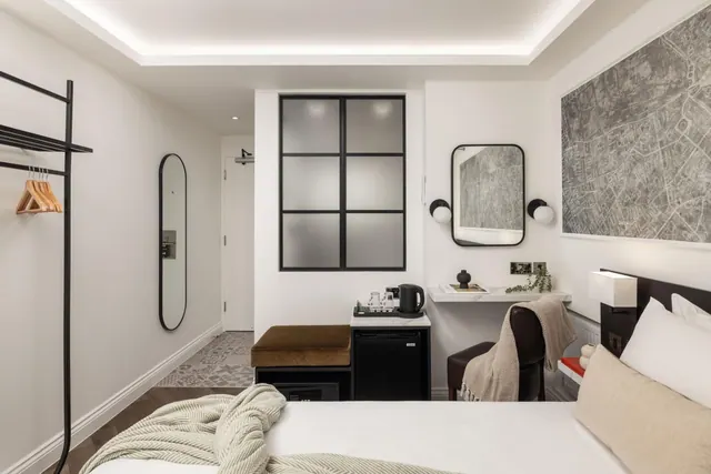 A hotel bedroom with a made double bed, a wall-mounted desk with a round mirror and chair, a kettle and tea tray on a minibar, a bench, and a free-standing clothes rail.