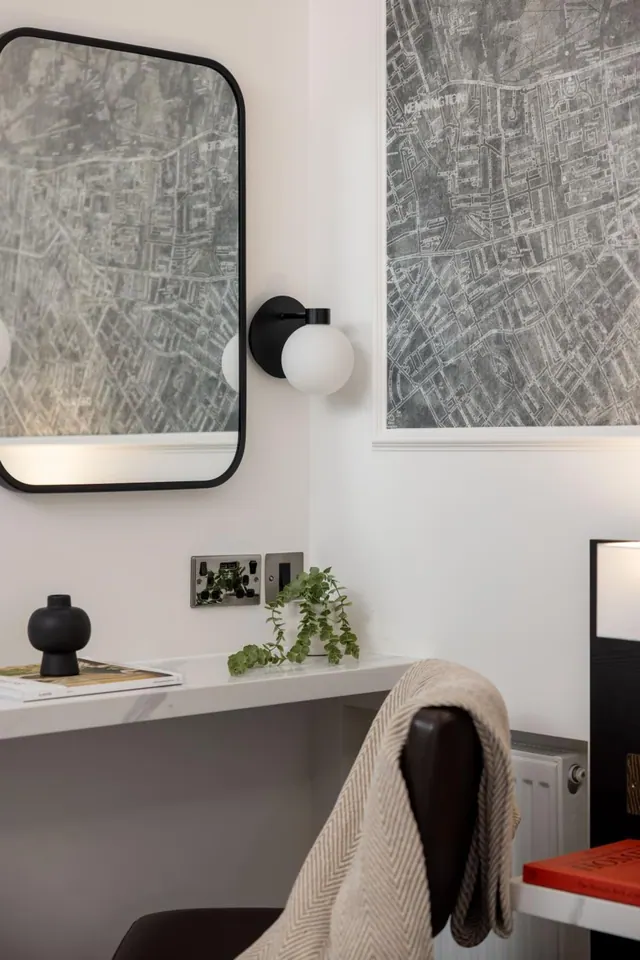 A corner of a room shows a wall-mounted mirror, a round wall light, a framed map print, a white wall shelf with a small plant and a black vase, and a chair draped with a beige throw.