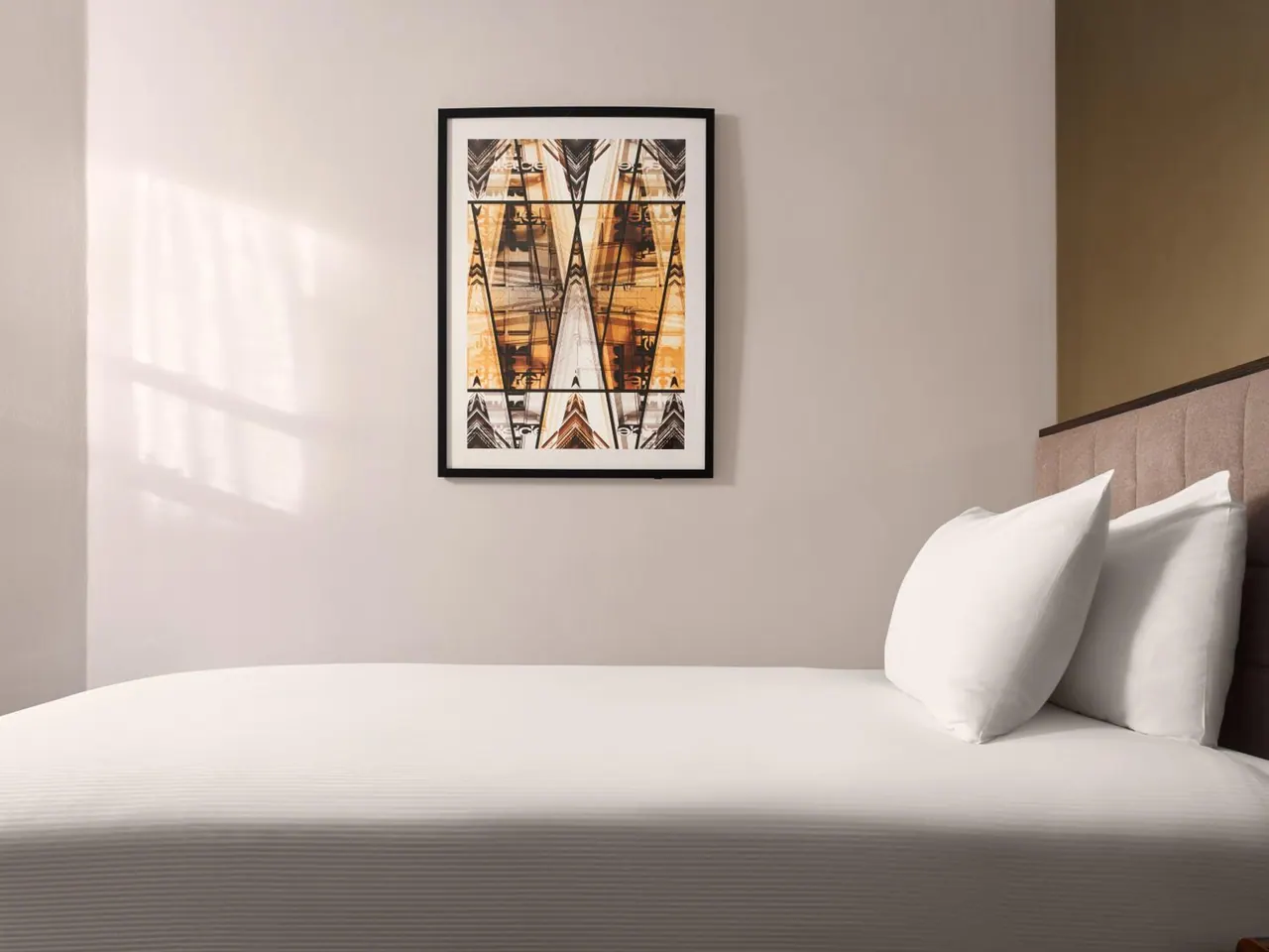 A made bed with white pillows and an upholstered headboard sits below a framed abstract print on a pale wall, with sunlight falling across the wall on the left.