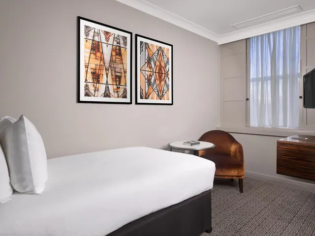 A bedroom with a made double bed, two framed abstract prints, a velvet armchair beside a small round table, a wall-mounted television and a window with sheer curtains.