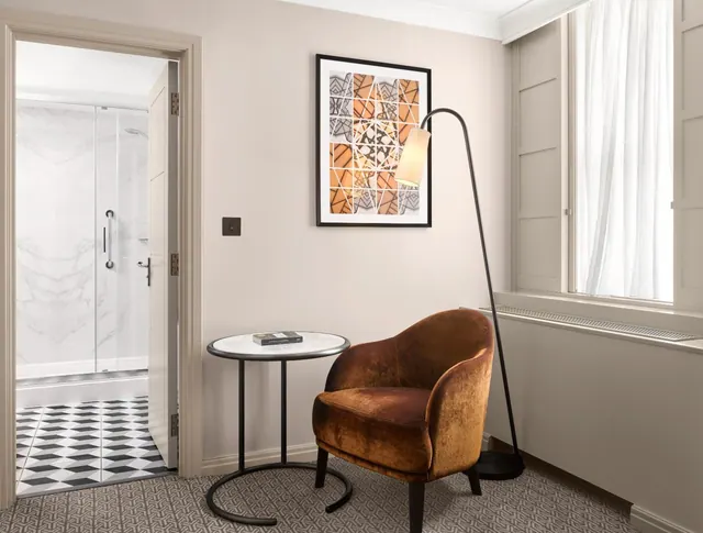 A room corner with an upholstered armchair, round side table, floor lamp and framed artwork, beside an open doorway to a bathroom with a walk-in glass shower.