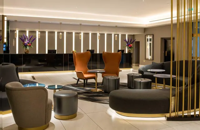 A hotel lobby with a reception desk along the back wall, orange armchairs, grey sofas and ottomans around low tables, and a gold rod divider on the right.