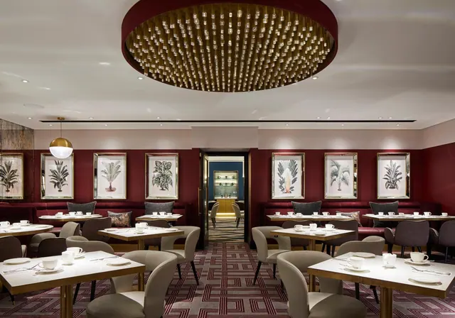 An empty restaurant dining room with set tables, upholstered chairs, burgundy banquette seating, framed botanical prints and a large circular ceiling light.
