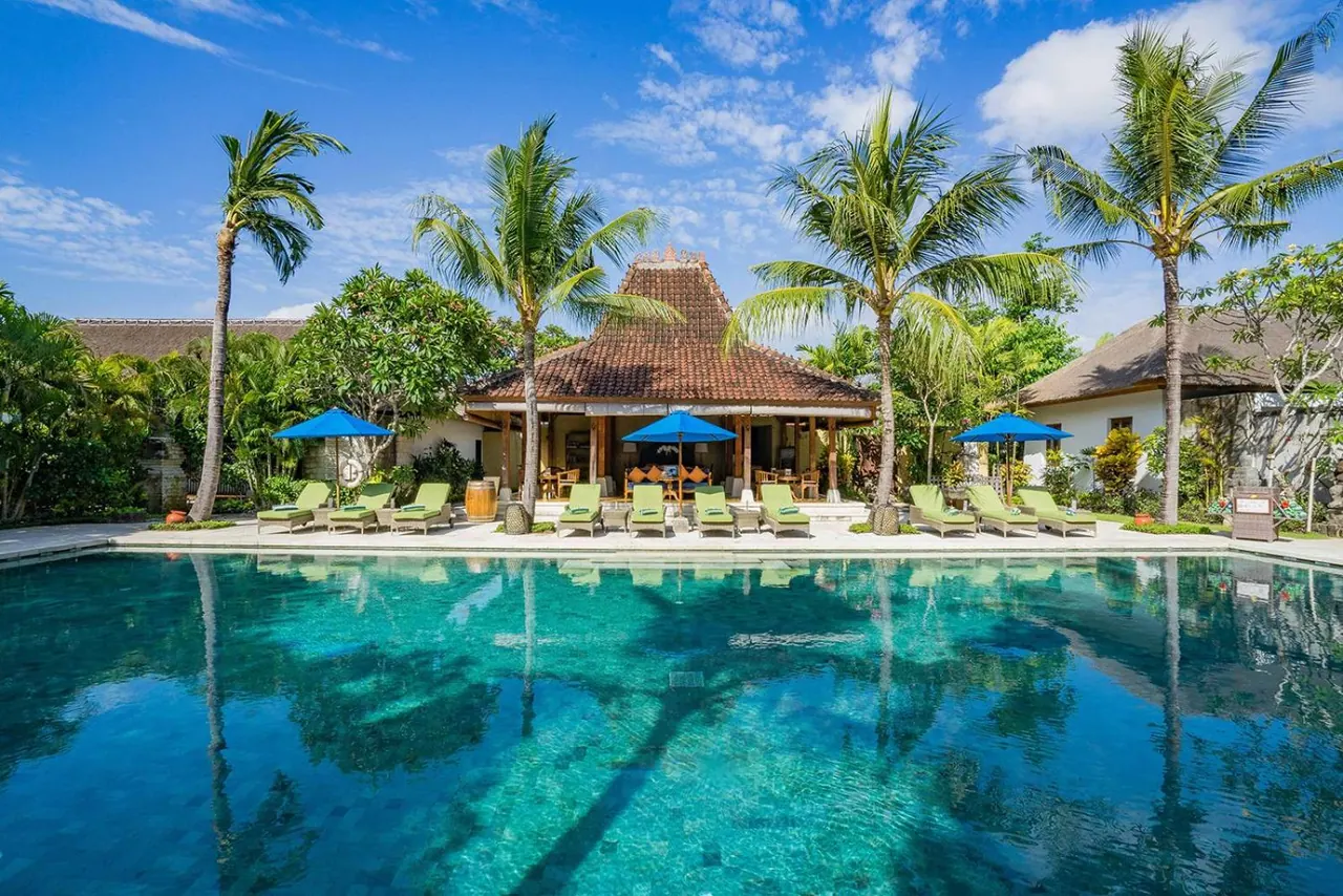 A swimming pool with green sun loungers and blue umbrellas sits in front of a traditional Balinese pavilion with palm trees under a clear blue sky.