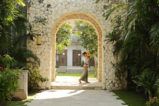 A woman in a sarong carrying an offering walks through a stone archway into a garden with a white building beyond.