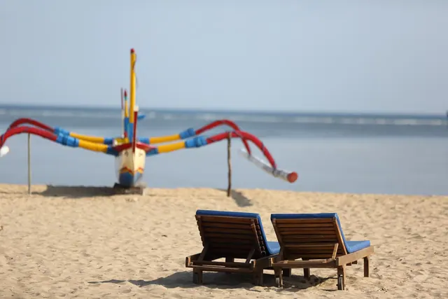 Two wooden sun loungers with blue cushions stand on sand in front of a traditional outrigger boat and calm sea.