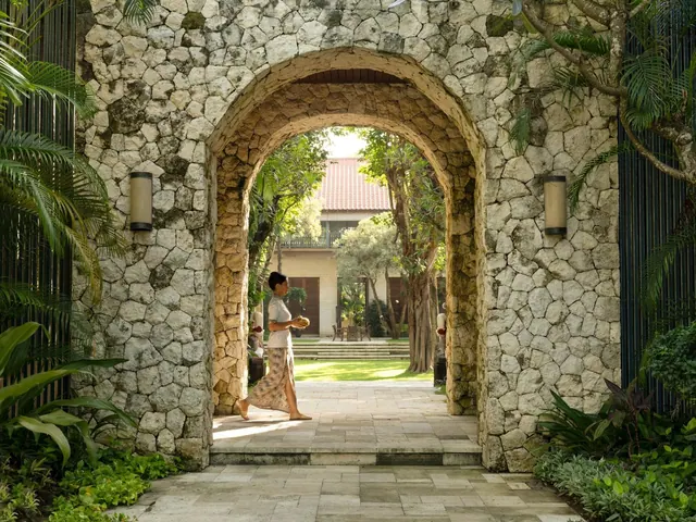 A woman in a sarong carries a fruit while walking through a stone arched gateway toward a lawn, trees and a building with a red tile roof.