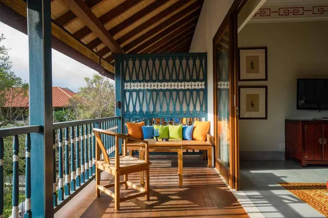 A covered veranda with a teak bench with coloured cushions, a teak chair and a blue railing, with an adjoining room containing a wall-mounted television and a wooden cabinet.