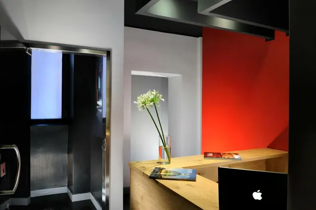 A light wood reception desk with a vase of white flowers, two magazines and a black monitor sits in front of a red wall, beside a lift with a steel frame.