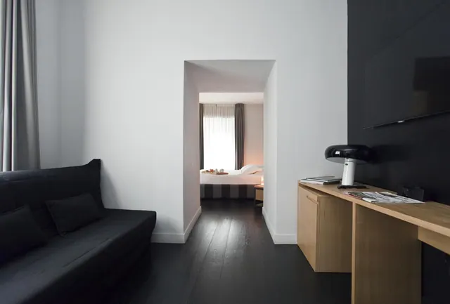 A black sofa sits in a living area beside a wall-mounted television and a wooden desk, with an archway leading to a bedroom containing a made bed and a sheer-curtained window.