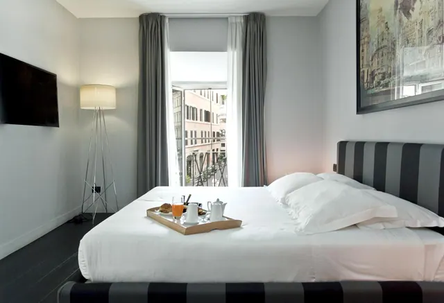 A made bed with a breakfast tray holding orange juice, a croissant and a tea service, beside a window looking onto neighbouring buildings.
