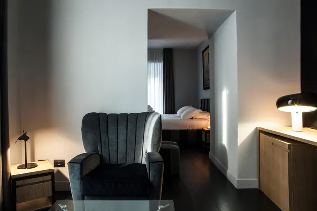 A grey armchair and glass coffee table sit in a room, with a made bed visible through an archway beyond.