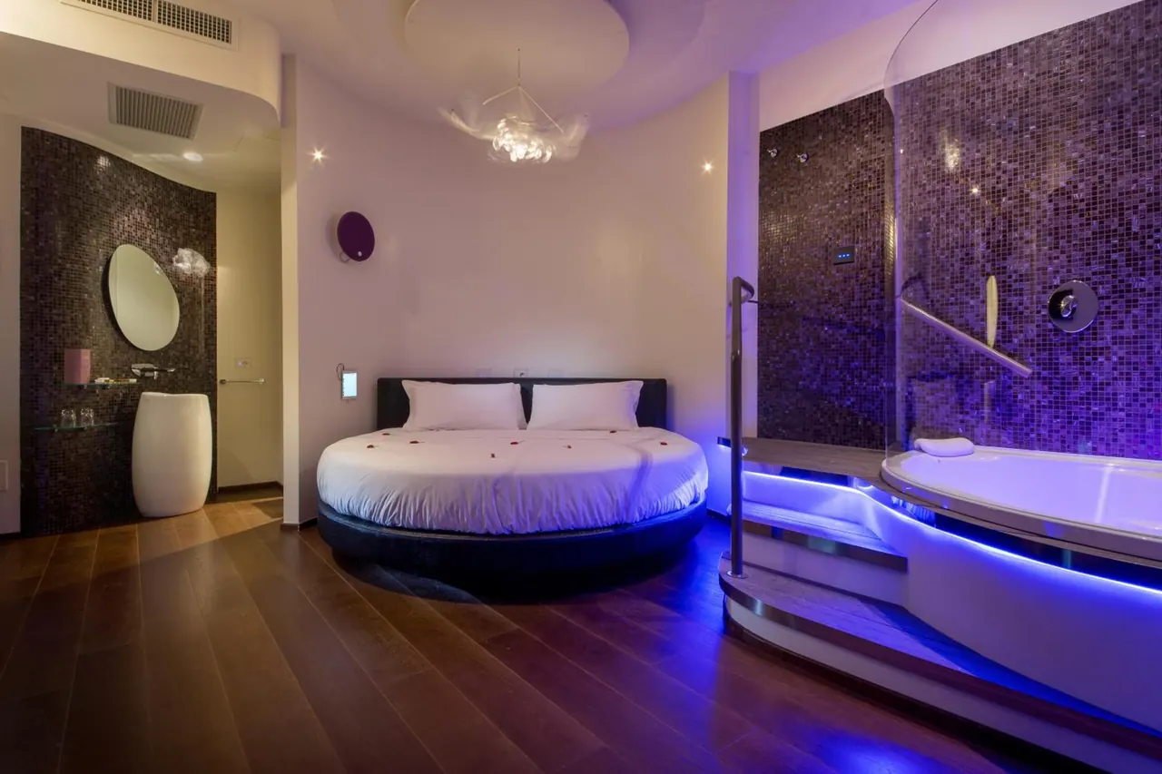 A bedroom with a round bed under purple lighting, a bathtub on a raised platform, and a pedestal basin beside a mosaic-tiled wall.