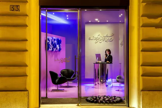 Night view through the glass entrance doors of a hotel reception area with purple lighting, a receptionist at a desk, and black swan chairs.
