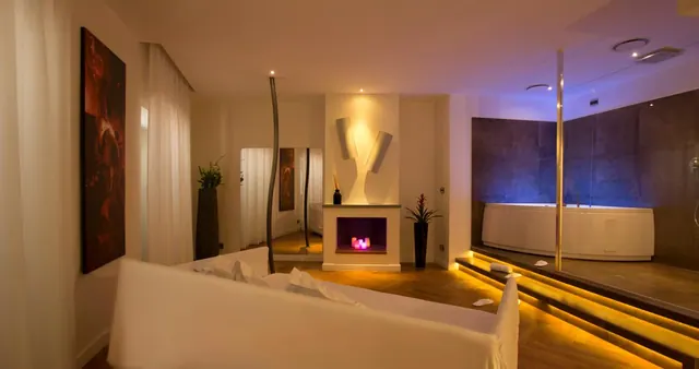 A suite living area with a white covered sofa, a fireplace niche with lit candles, and a bathtub on a raised platform with LED step lighting.