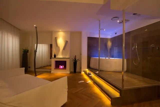 A bedroom with a large white bed, a glass-walled walk-in shower and a freestanding bathtub on a raised platform, lit by warm and blue lights.