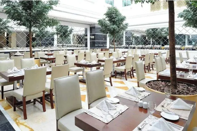 A dining room with cream leather chairs and wooden tables set with folded napkins and glassware, with indoor trees growing under a bright atrium.