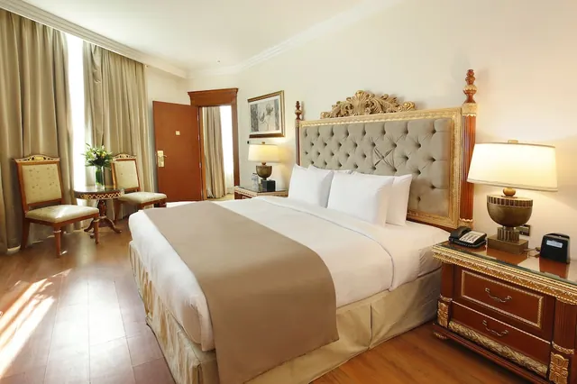 A bedroom with a large bed under a tufted grey headboard with gold trim, two upholstered chairs beside a small round table, and a wooden floor.