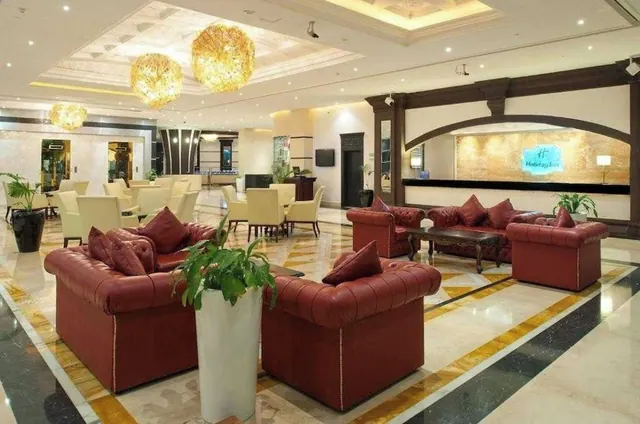 A hotel lobby with red leather chesterfield sofas, ornate gold chandeliers, a marble floor, and a lit Holiday Inn logo on the reception wall.