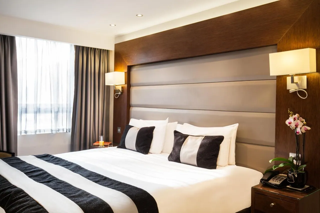 A made-up king bed with a black and white striped runner and cushions, with wall lamps, a telephone and an orchid on the bedside table beside a window with curtains.