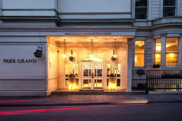Front entrance of the Park Grand Paddington Court hotel at dusk, with illuminated glass doors, lanterns and hanging planters on a white stucco facade.