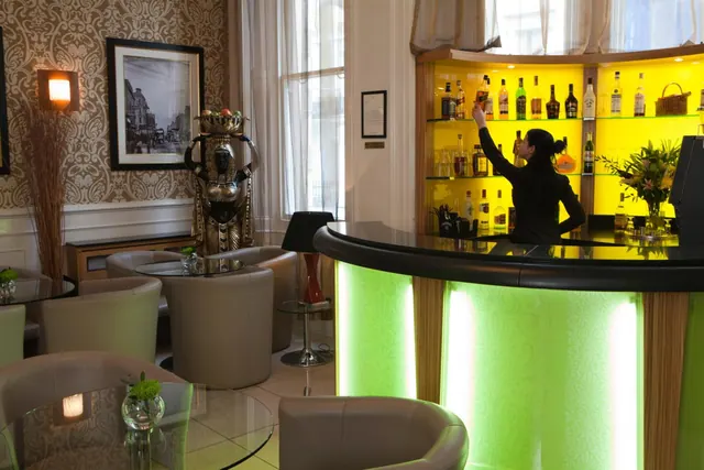 A bartender reaches for a bottle behind a curved bar with green-lit panels and yellow-backlit shelves, with armchairs and glass tables in the foreground.