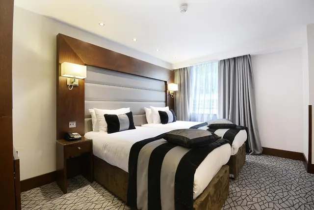 A hotel bedroom with twin beds dressed in white linen and black and grey striped runners, with a bedside telephone and a curtained window.