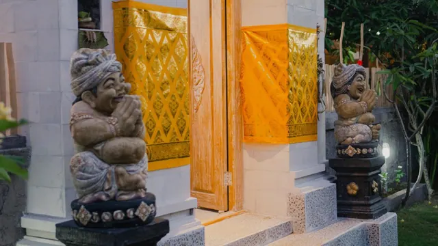 Two stone guardian statues sit on pedestals beside a wooden door, with gold cloth draped over the white pillars of a Balinese-style entrance.