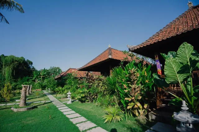 A stone-paved path runs across a lawn toward traditional Balinese pavilions with terracotta-tiled roofs, with tropical plants and stone lanterns beside it.