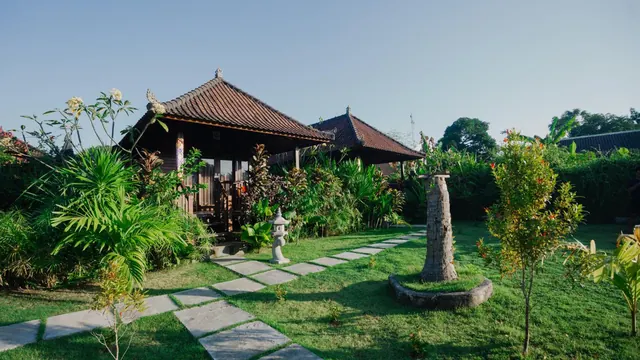 A garden with a traditional Balinese pavilion, a stone paved path across the lawn, a stone lantern and a carved tree-trunk sculpture on a grass island.