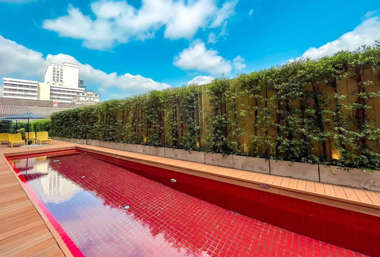 A long rooftop pool with red mosaic tiles sits beside a timber deck, a tall green plant wall, and yellow sun loungers, with city buildings visible at the left under a blue sky.