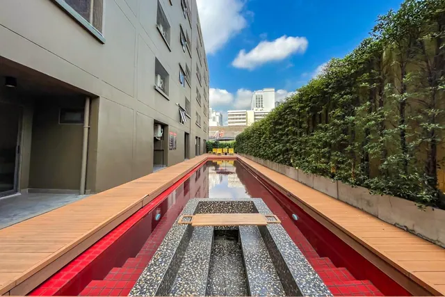 A long narrow red-lined water channel with a terrazzo crossing and wooden bench runs between a building wall and a tall green hedge, with city buildings visible beyond.