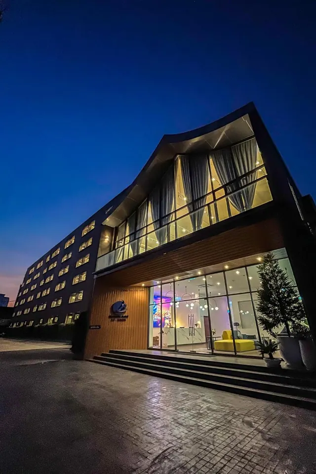 Exterior of a modern hotel building at dusk, with a glass-fronted entrance, wide steps and illuminated windows.