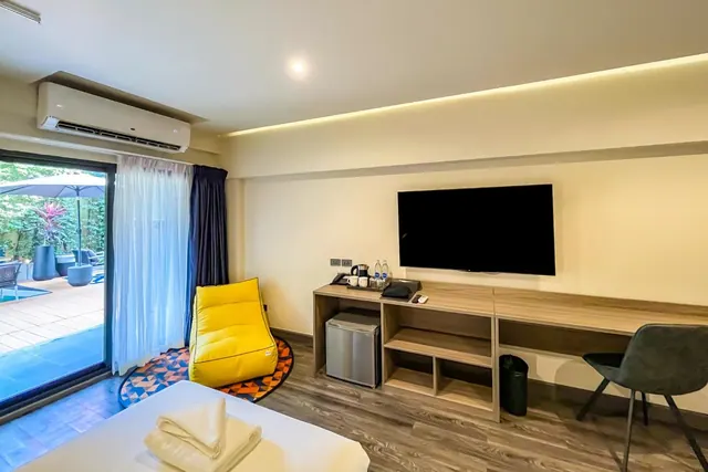 A hotel room with a glass sliding door onto an outdoor terrace and pool, a yellow bean bag chair, a wall-mounted television, a wooden desk with a kettle and a small fridge beneath.