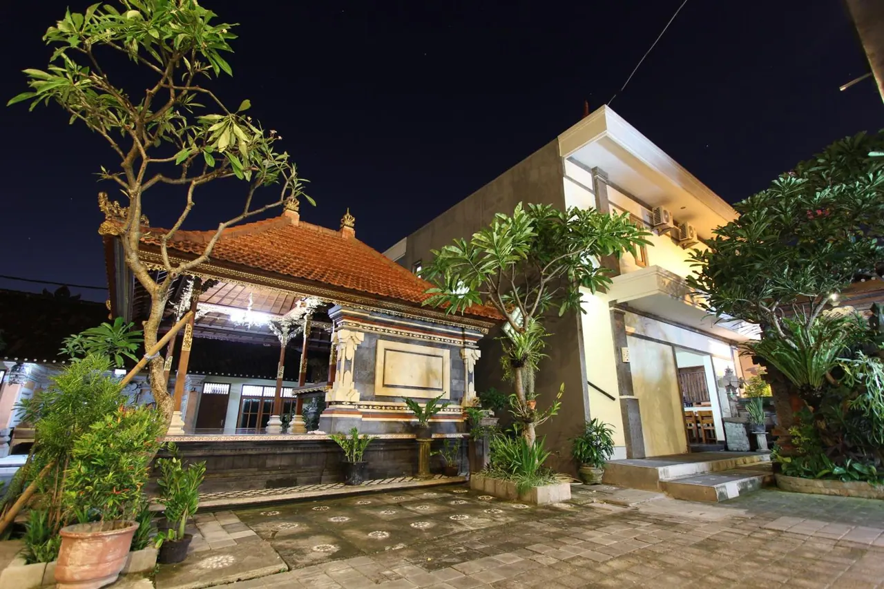 A night view of a traditional Balinese pavilion with a terracotta roof and frangipani trees, beside a multi-storey building with a paved courtyard in front.