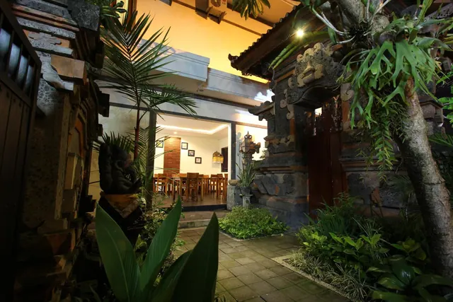 A night view of a planted paved courtyard path passing a carved stone Balinese gate, with an indoor dining room with wooden tables and chairs visible beyond.