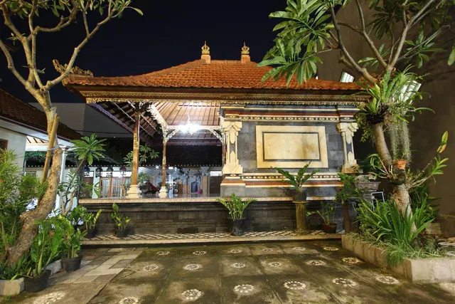 A lit traditional Balinese pavilion with a tiled roof stands in a courtyard at night, flanked by frangipani trees and potted plants.